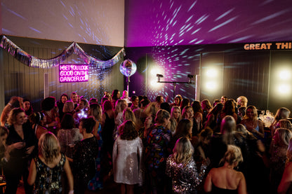 A crowded indoor event space with many people dancing under purple and pink disco lighting, a neon sign reading 'I BET YOU LOOK GOOD ON THE DANCEFLOOR,' 