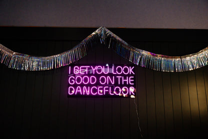 A pink neon sign on a dark paneled wall that reads 'I BET YOU LOOK GOOD ON THE DANCEFLOOR', decorated with a shiny silver tinsel garland above it.
